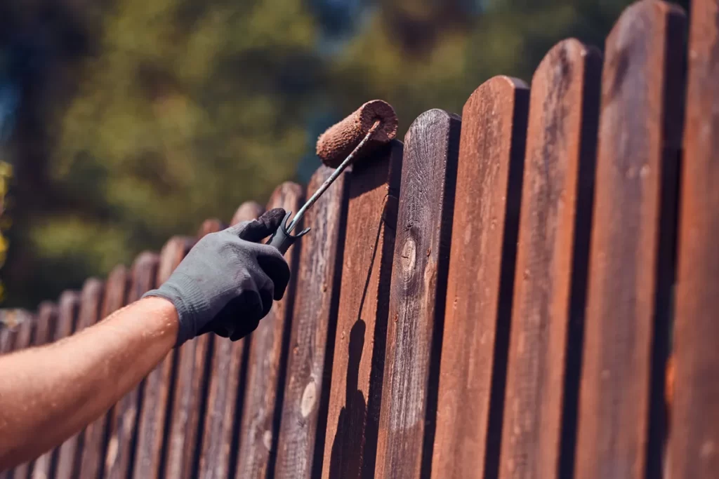 man protective gloves is painting wooden fence bright summer day